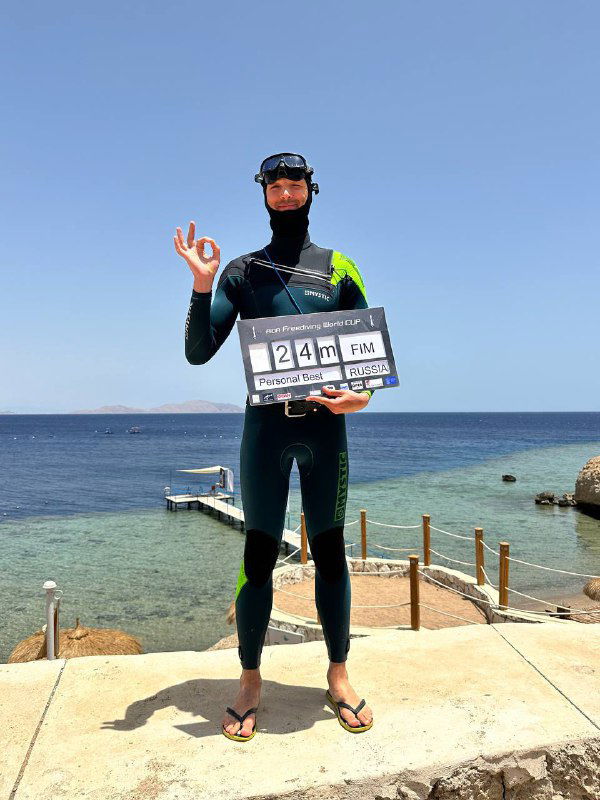 Фридайвер в гидрокостюме и маске держит табло с цифрой 24 м, позирует на каменном берегу у синего моря после успешного нырка в лагере.