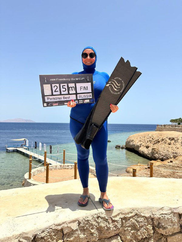 Фридайвер в синим гидрокостюме держит ласту и табло с результатом 25 м, стоит на фоне моря и скал, солнечная площадка для старта и фото.