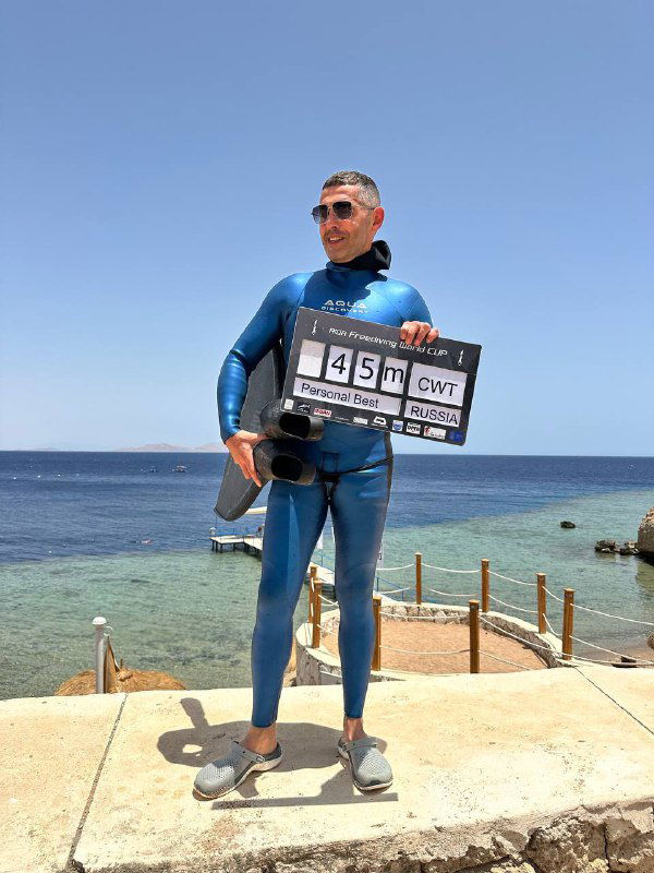 Фридайвер-мужчина стоит на побережье с табло 45 м в руках и снаряжением, на фоне море и причал; момент после успешного глубокого погружения.