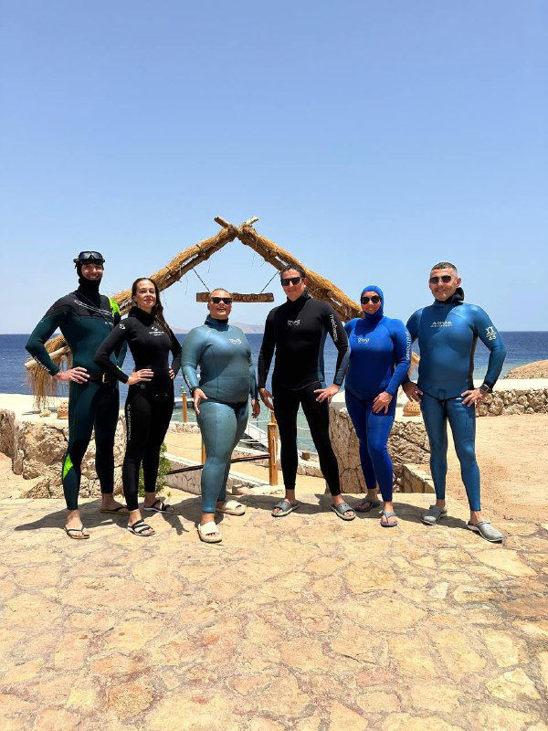 Группа участников в гидрокостюмах собралась у каменной арки на берегу, совместное фото команды фридайверов после завершения тренировок в Шарме.