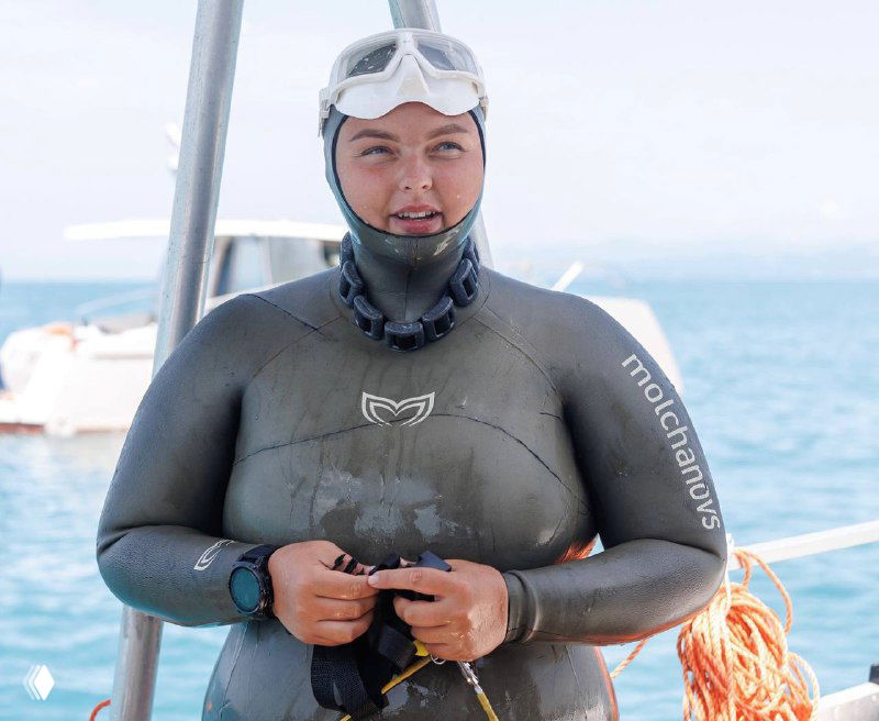 Портрет фридайвера в гидрокостюме на палубе: маска, неоприновый костюм и часы для контроля погружения на фоне моря.