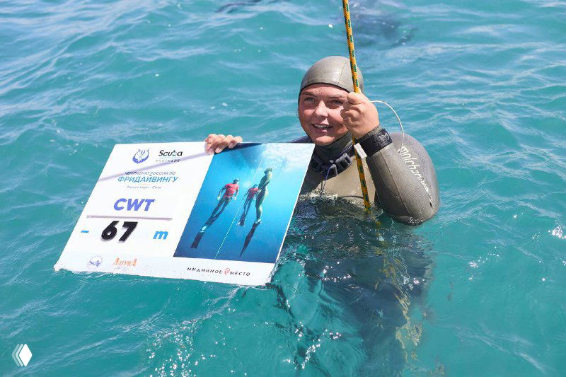 Фридайвер держит табличку с номером и результатом на воде, вокруг видна команда и снаряжение, морской фон соревнований.