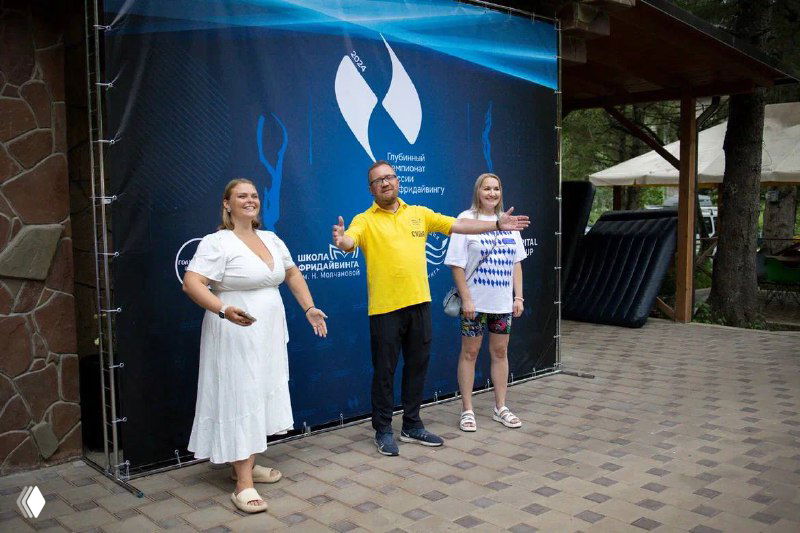 Три человека у баннера чемпионата: организатор в жёлтой рубашке и участница в белом приветствуют публику перед началом соревнований по фридайвингу.