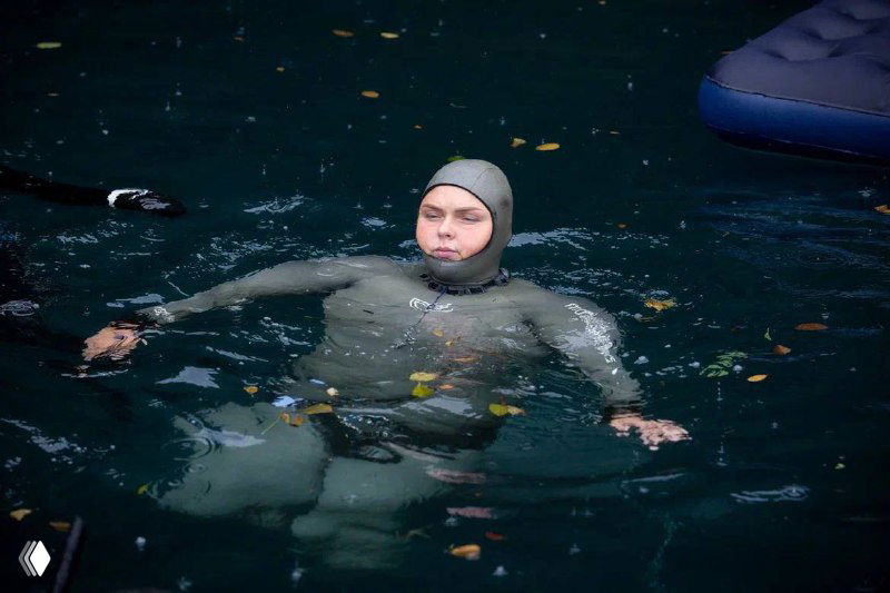 Фридайвер в гидрокостюме плавает на поверхности тёмной воды во время соревнований, вокруг видны листья и спокойная поверхность водоёма.