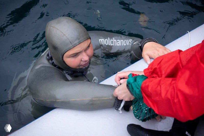 Фридайвер в гидрокостюме тянется к борту или лодке, помощник протягивает руку — момент подъёма после задержки дыхания на соревнованиях.