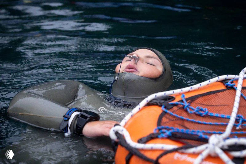 Фридайвер отдыхает на поверхности воды после задержки дыхания, лицо расслаблено, команда наблюдает за восстановлением перед следующим стартом.