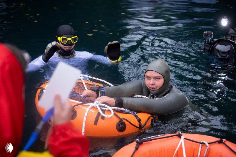 Фридайверы у берега получают инструкции и записи от судьи; участник в гидрокостюме держится за плот, фиксируются результаты и подписи.