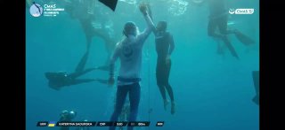 Подводный кадр соревнований по фридайвингу: спортсменка всплывает к поверхности, рядом судьи и участники на фоне синей воды, кадр из видео.