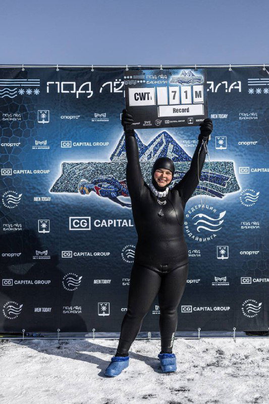 Ольга Маркина поднимает табло с отметкой «71 м» на фоне баннера соревнования «под лёд Байкала», улыбка и логотипы спонсоров видны отчётливо.