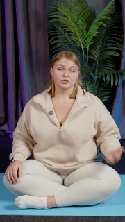 Ольга Маркина сидит на коврике в студии и показывает технику диафрагмального дыхания; позади — шторы и зеленое растение.