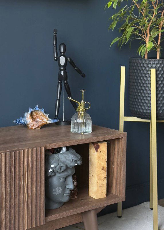 Декор на тумбе: фигурная статуэтка, ракушка и стеклянный распылитель на фоне тёмно-синей стены и зелёного растения в интерьере.