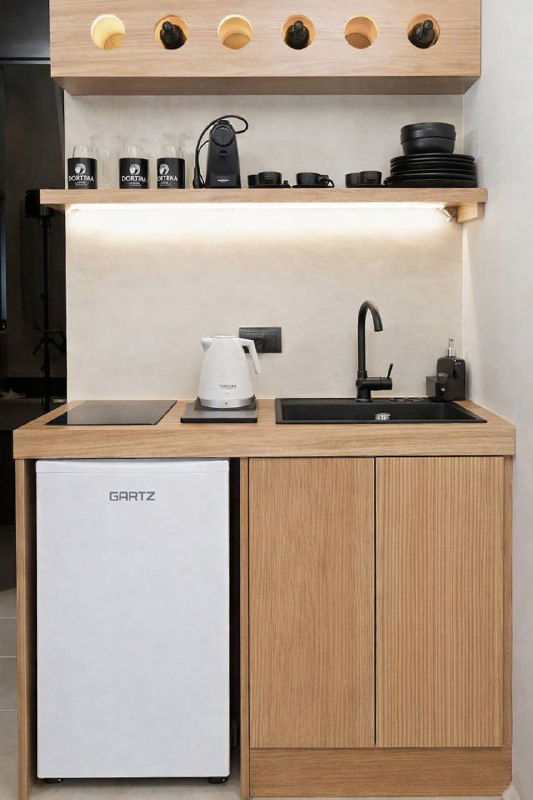 Белый мини‑холодильник GARTZ в компактной кухне с деревянным гарнитуром: раковина, чайник на столешнице, открытые полки и подсветка.