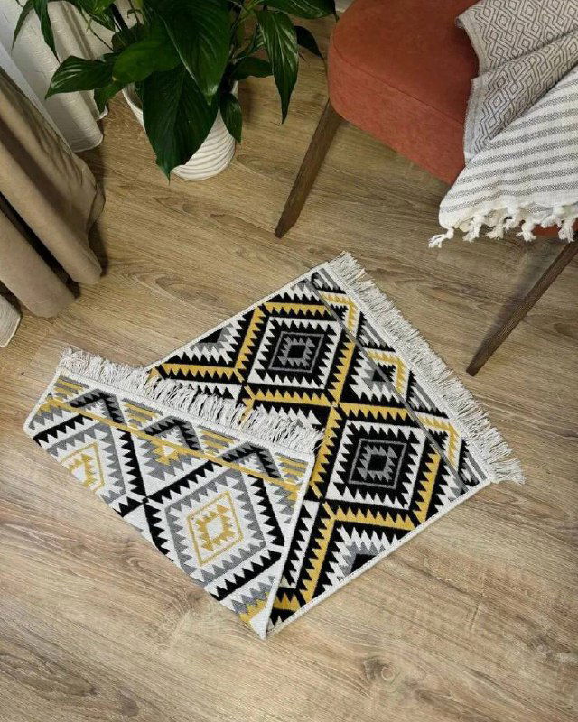 Прикроватный коврик с геометрическим чёрно‑белым узором и жёлтыми акцентами на деревянном полу рядом со стулом и растением