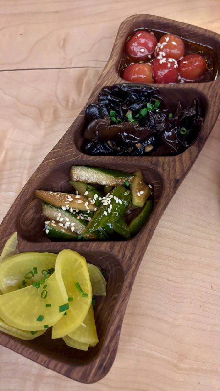 Деревянная разделённая тарелка с четырьмя типами цукэмоно: маринованный дайкон, огурцы, грибы и томаты, украшенная кунжутом и зеленью.