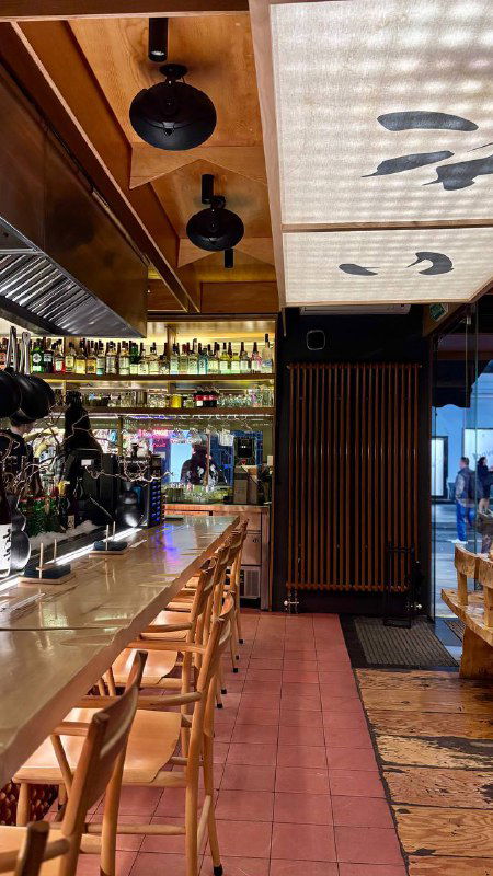 Интерьер ресторана: длинная барная стойка, высокие стулья и японская вывеска — атмосферное вечернее пространство заведения.