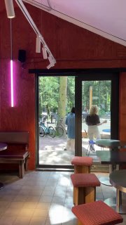 Интерьер кафе с видом на выход в лес: барные стулья, большие двери и прохожие у велосипедов на летней площадке за стеклом.