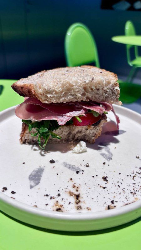 Крупный план сэндвича с прошутто на цельнозерновой булочке на белой тарелке, видно листья салата и ломтики прошутто на зелёном столе.