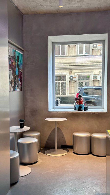 Уютный уголок у большого окна в кофейне 2046: столики, пуфы и вид на улицу через окно, художественные предметы на стене.