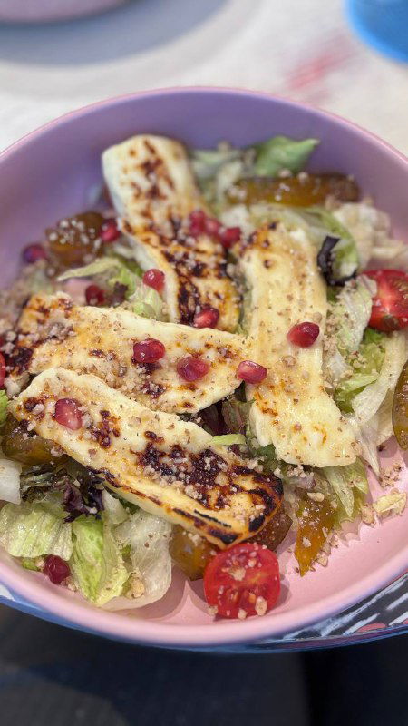 Салат с жареным халуми: полоски сыра на листах салата, сверху зерна граната и черри, в розовой тарелке на цветном столе.