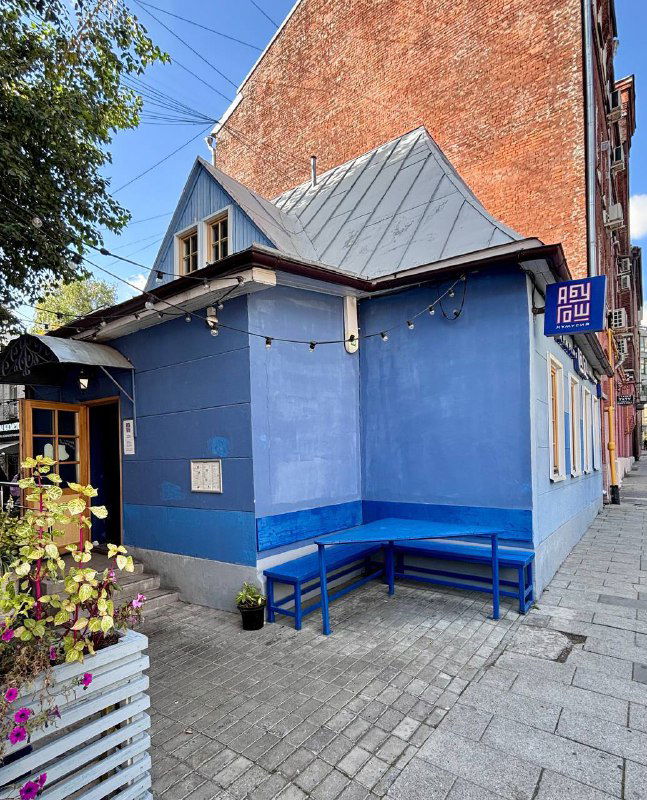 Фасад кафе в синей гамме с скамейкой у входа: небольшой угловой дом и вывеска сети «Абу Гош», городской пейзаж Москвы на фоне.