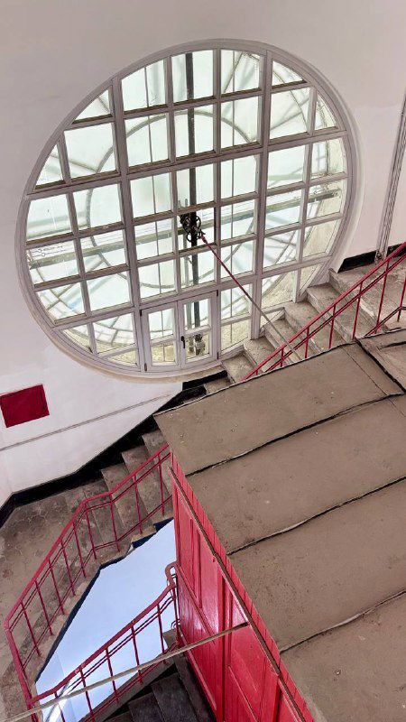 Овальное световое окно и металлическая лестница в шахте башни: вид вниз по лестнице, красные перила и конструкция смотровой площадки у часов.