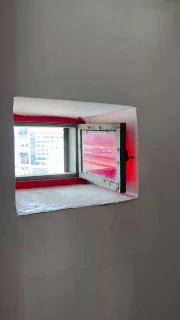 Небольшое квадратное окно с красной рамой в комнате башни, вид на внутреннее пространство и фрагмент стены с покраской и вентиляционным проемом.