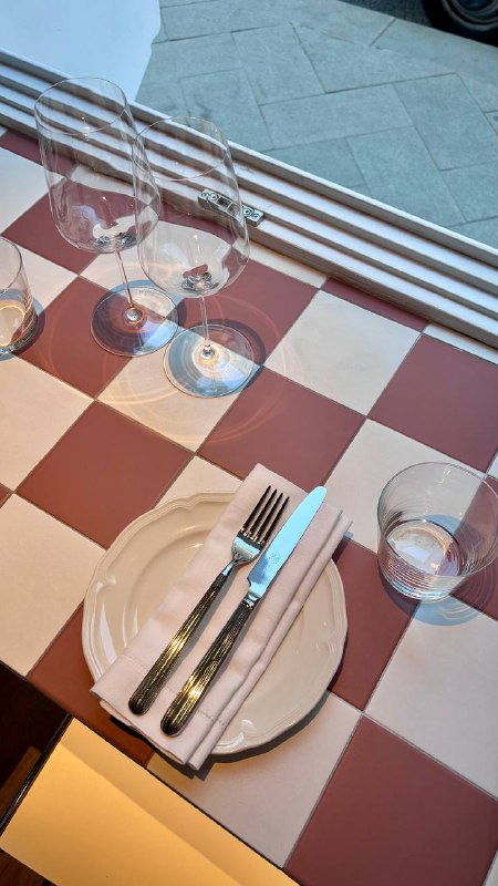 Сервировка на столе у окна: стеклянная посуда, приборы и клетчатая скатерть; дневной свет создаёт уютную атмосферу ресторана