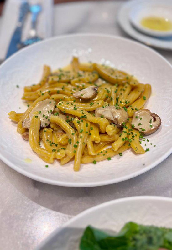 Пастообразное блюдо с трубчатыми макаронами в сливочном соусе и грибами, поданная на белой глубокой тарелке на столе.