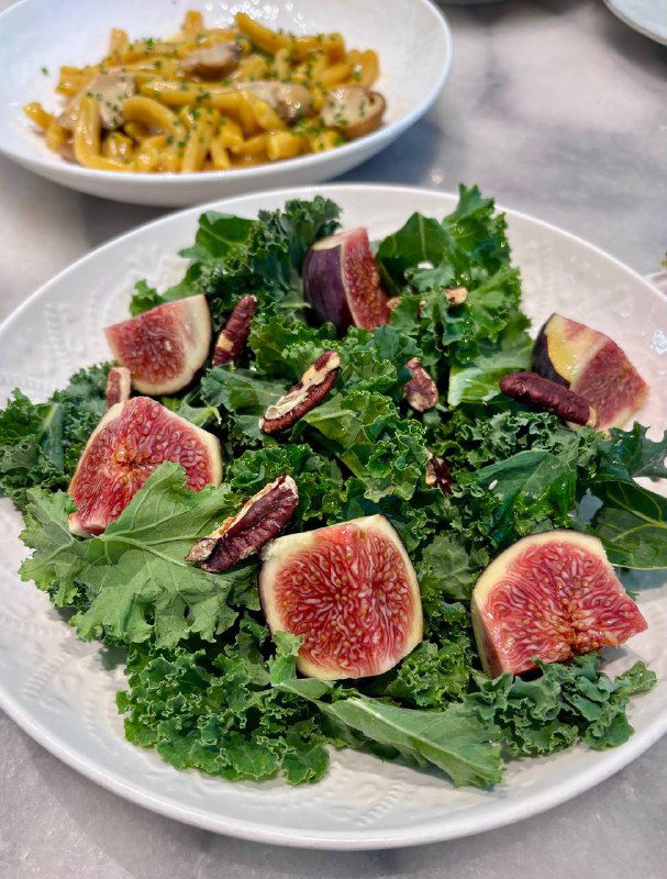 Большой салат с листовой зеленью, дольками свежих инжиров и орехами на белой плоской тарелке — акцент на сезонность.