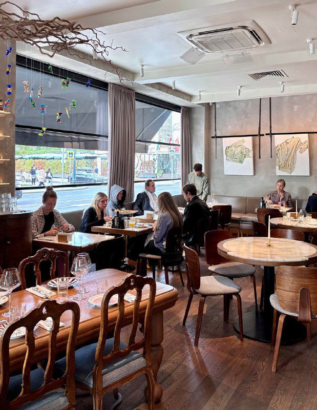 Интерьер Linbistro: зал с деревянными столами и стульями, большими окнами и естественным светом, вид на посетителей.