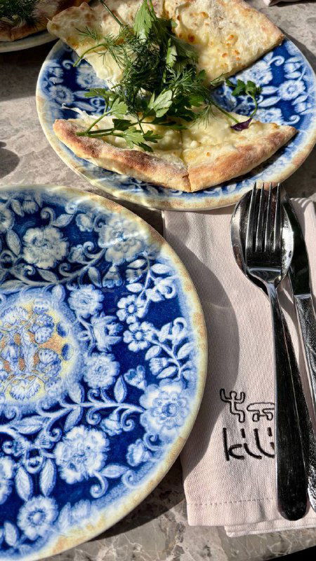 Лепёшка с зеленью на синей керамической тарелке и фирменная салфетка с логотипом Kilim на столе — деталь сервировки и брендинга ресторана.