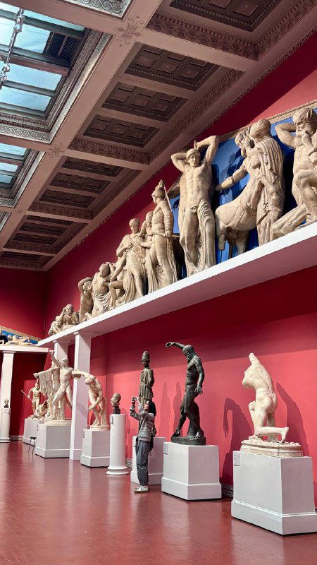 Ряд классических статуй на постаментах вдоль стены с высоким потолком и большим световым люком; ракурс подчёркивает масштаб и ритм экспозиции.