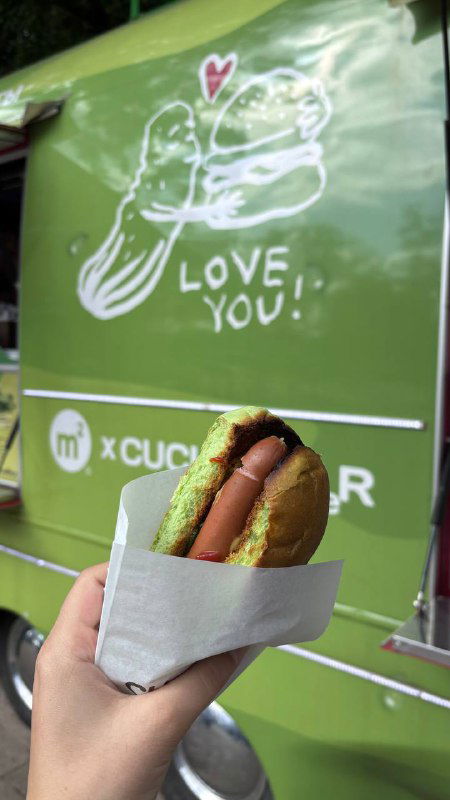 Крупный план хот-дога с зелёной булочкой в бумажной обёртке на фоне ярко-зелёного фудтрака с рисунком и надписью.