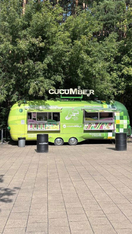 Ярко-зелёный фудтрак "Cucumber" на солнечной полянке в Мещерском парке, припаркован на фоне высоких деревьев.