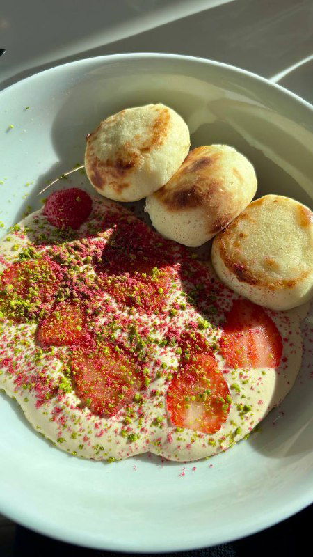 Тарелка с сырниками и ягодами: сверху воздушный крем из сгущенки, посыпка и кусочки инжира рядом, солнце падает через окно кафе.