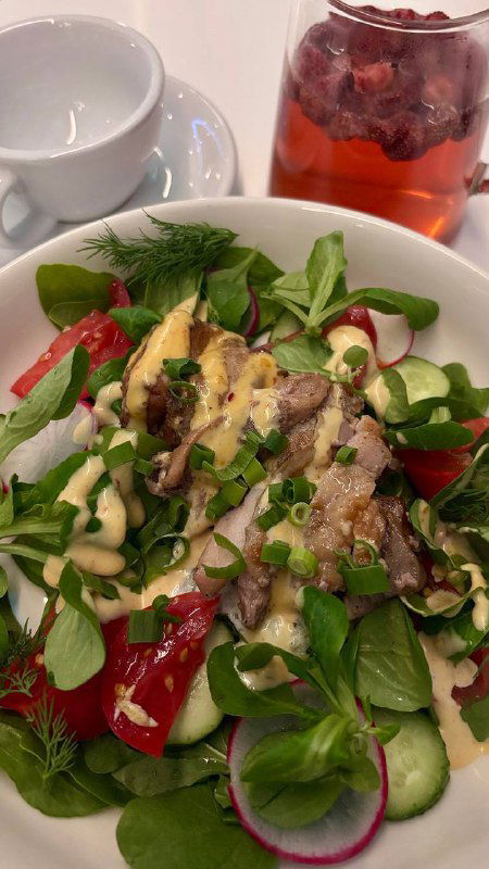 Большая тарелка салата с миксом зелени, ломтиками овощей и заправкой; на заднем плане стакан с травяным чаем цвета ягод.