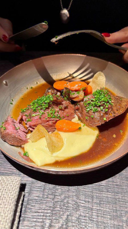 Тарелка со средне-прожаренным мясом, овощами и соусом на столе; вид с бокового ракурса, столовые приборы и часть сервировки в кадре.