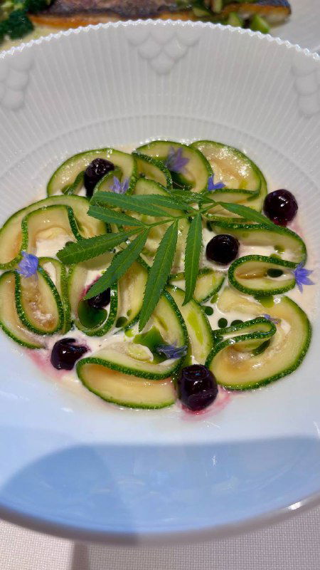 Тонко нарезанные цукини с зеленью и точками чёрной смородины на тарелке — лёгкая закуска со страчателлой и свежими акцентами.