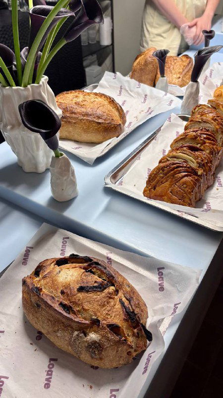 Ещё несколько буханок и выпечка разложены на столе в пекарне, видны корочки, текстуры и упаковочная бумага с брендом Loave.