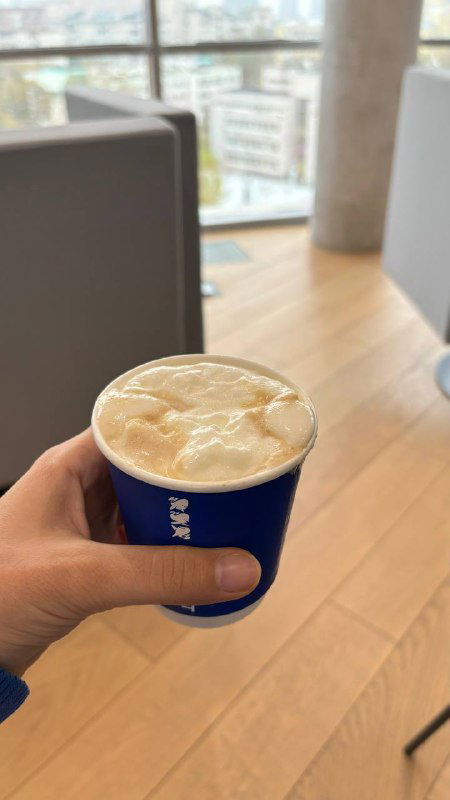 Рука держит бумажный стакан с кофе и плотной творожно-сырной пенкой на фоне окна с видом на город и интерьер кофейни.