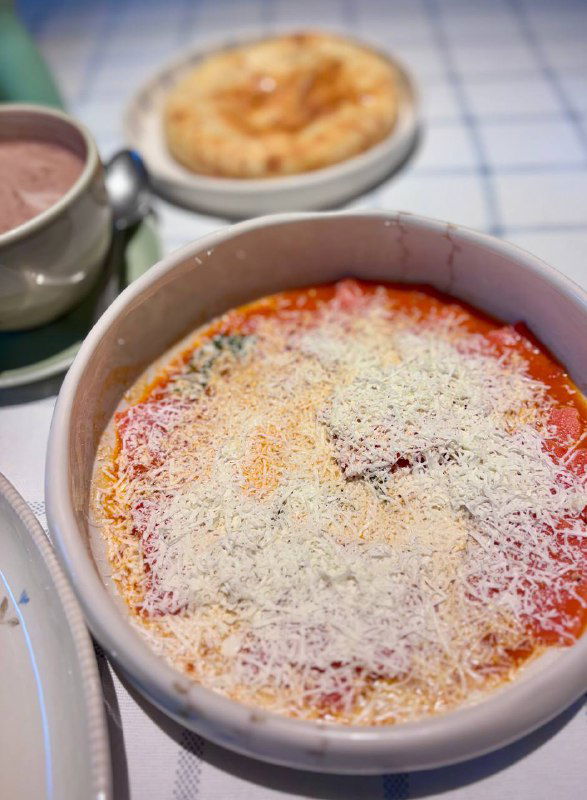 Глубокая миска с домашней пастой в томатном соусе, щедро посыпанная тёртым сыром, снята на столе с хлопковой скатертью.