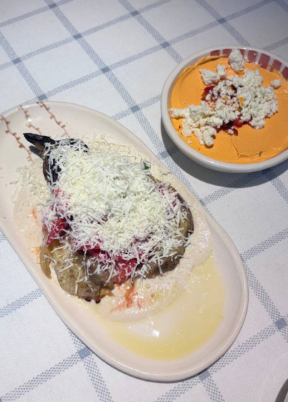 Запечённый баклажан с фетой, томатами и несколькими соусами, посыпанный тёртым сыром, кадр крупным планом на тарелке.