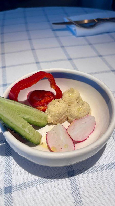 Маленькая миска с мезе: перец ромиро и фета, рядом свежие овощи и элементы закуски, подающейся как комплимент в ресторане.