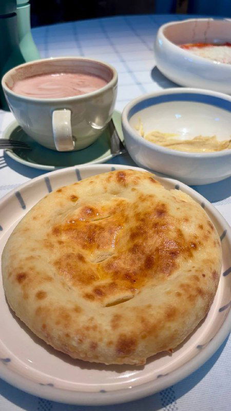 Круглая пита с золотистой корочкой на тарелке, рядом чашка напитка и соус — простая и съедобная хлебная основа блюда.