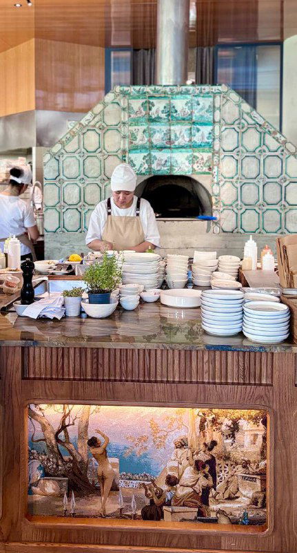 Открытая кухня и печь с орнаментальной плиткой, повар за работой и стойка с тарелками — видно кулинарный антураж заведения.