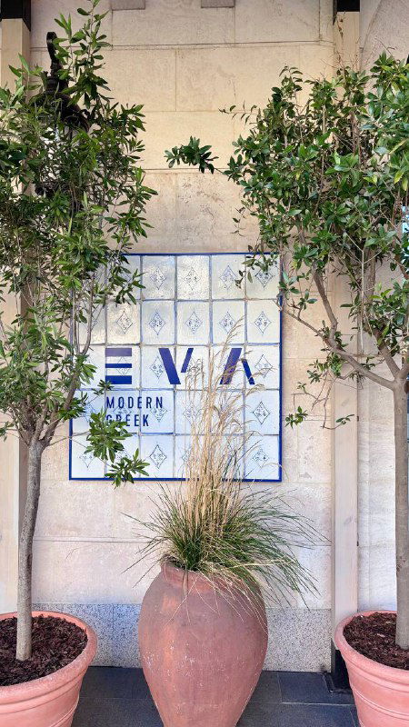 Фасад ресторана с плиточной вывеской "EVA" и горшками с растениями у входа, приветливый городской вход рядом с метро Киевская.