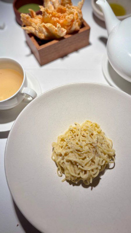 Миниатюрная порция тальялини на белой глубокой тарелке, рядом чашка с соусом и хрустящая закуска на заднем плане — минималистичная подача блюда.