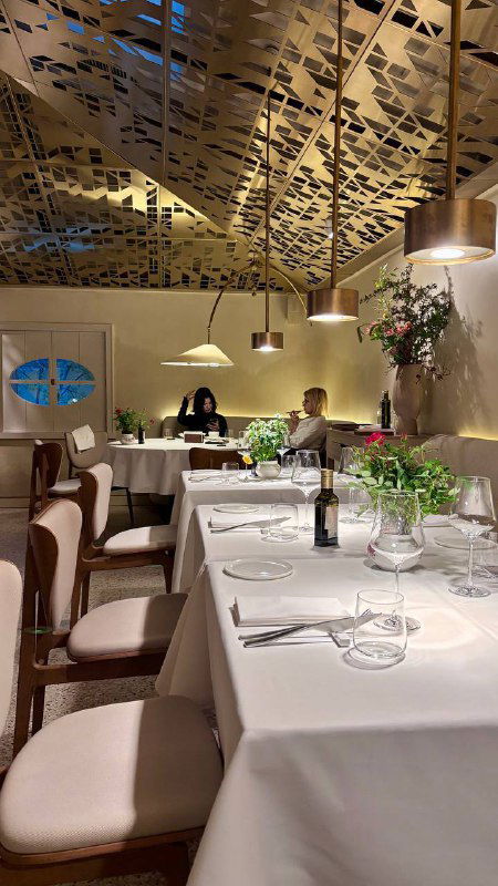 Зал ресторана с гостями за столиками, белые скатерти, зелёные растения и приглушённое освещение — общий вид атмосферы и обстановки.