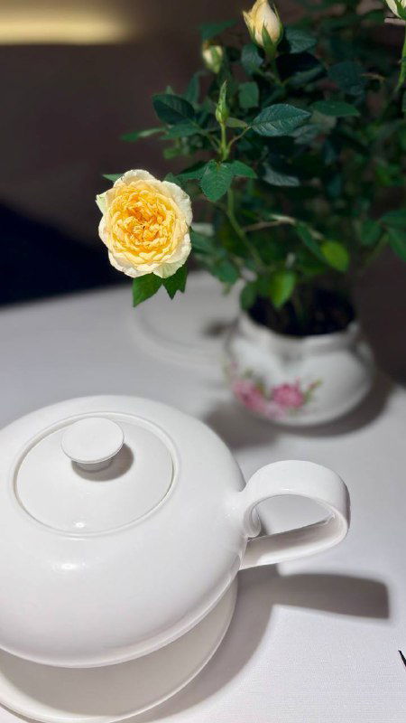 Жёлтая кустовая роза в небольшой фарфоровой вазе на столе рядом с белым чайником — элемент цветочной сервировки и декоративная деталь в зале.