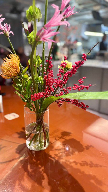 Ваза с высоким букетом цветов на столе внутри заведения; на фоне видна рабочая зона кухни и интерьер кафе с людьми.